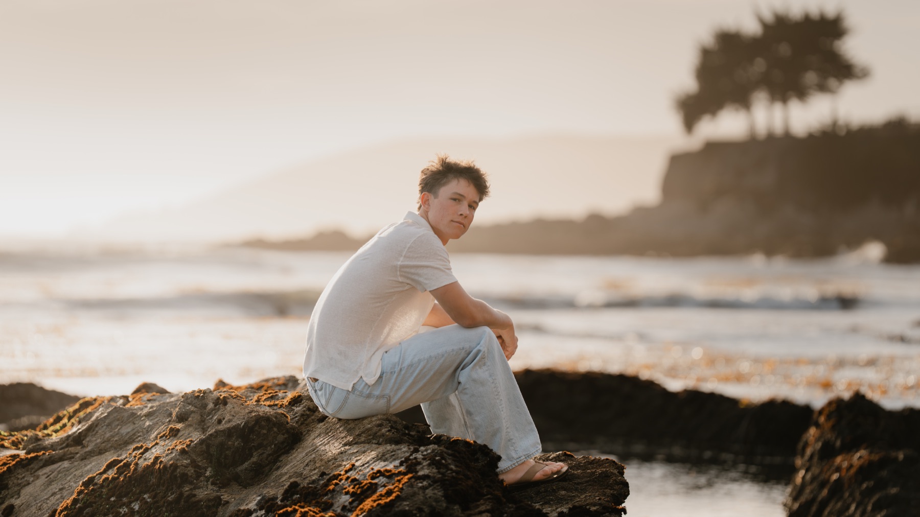 Senior sitting on coastal rocks at sunset, looking back at the camera — Central Coast senior photographer