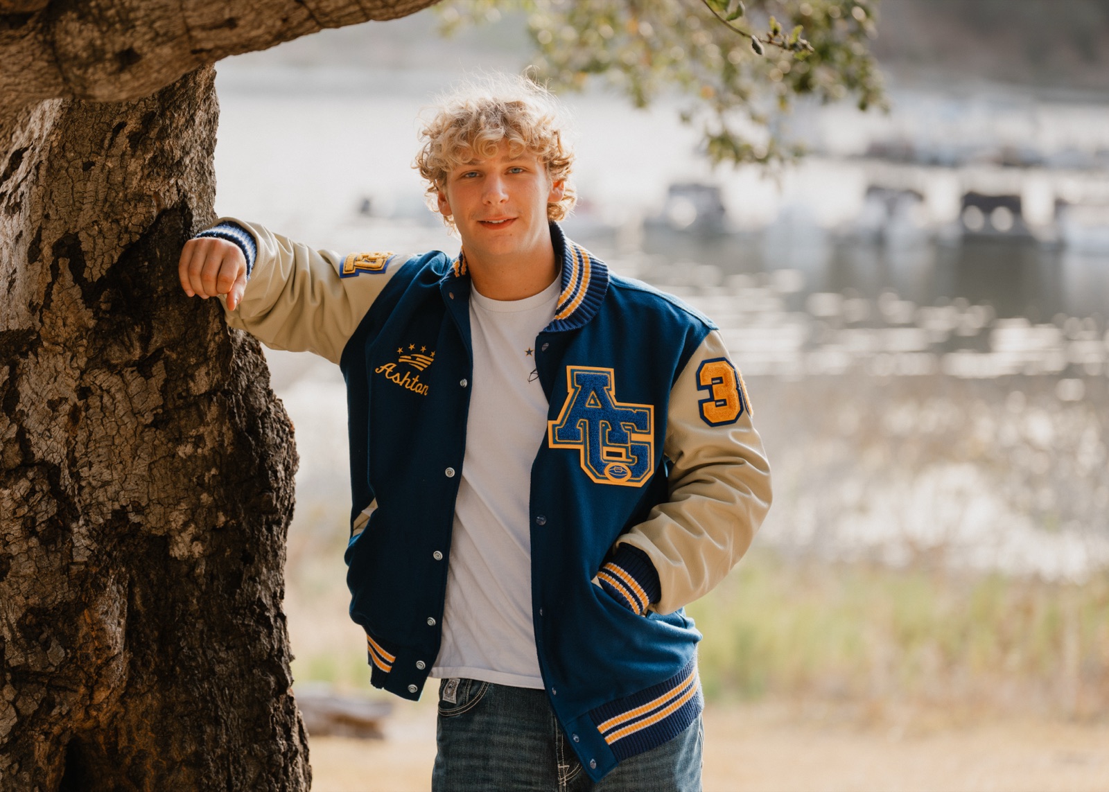 Senior in an Arroyo Grande varsity letterman jacket leaning on an oak tree by the lake — Arroyo Grande senior photographer