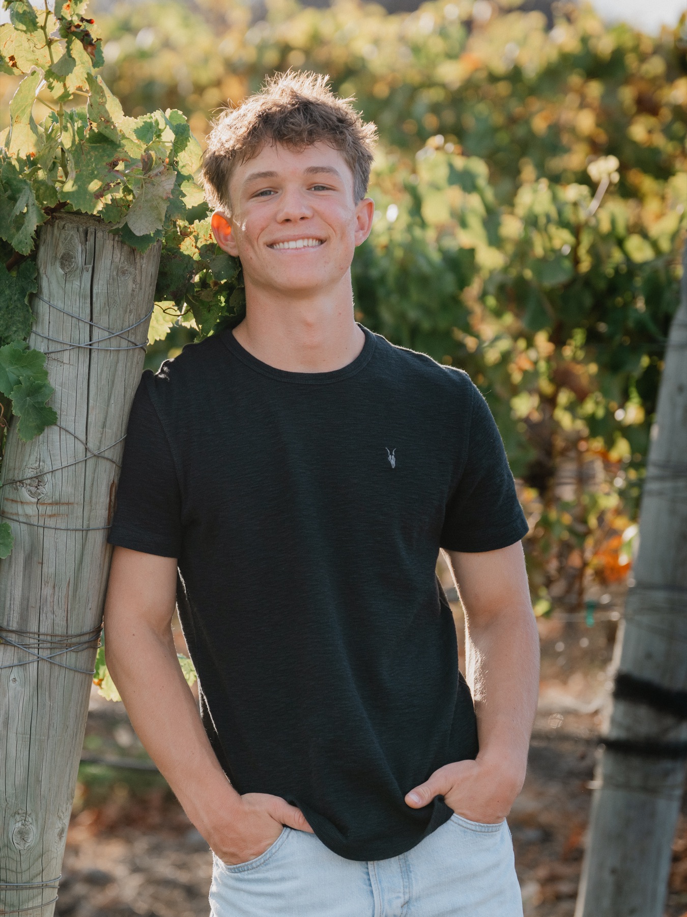 Senior smiling, leaning on a vineyard post in golden light — Central Coast senior photographer